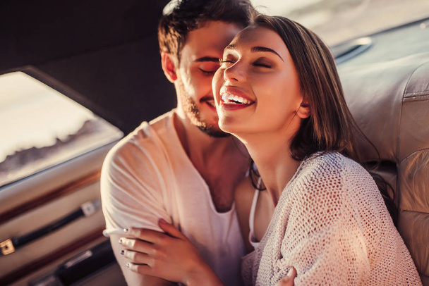 Romantic couple is sitting in green retro car on the beach. Handsome bearded man and attractive young woman in vintage classic car. Stylish love story. Hugging and kissing while being in car. - 写真・画像