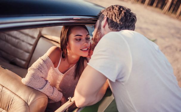 Romantic couple with green retro car on the beach. Handsome bearded man and attractive young woman with vintage classic car. Stylish girl is sitting in car while her man is standing near and is gonna kiss her. Love story. - Foto, imagen