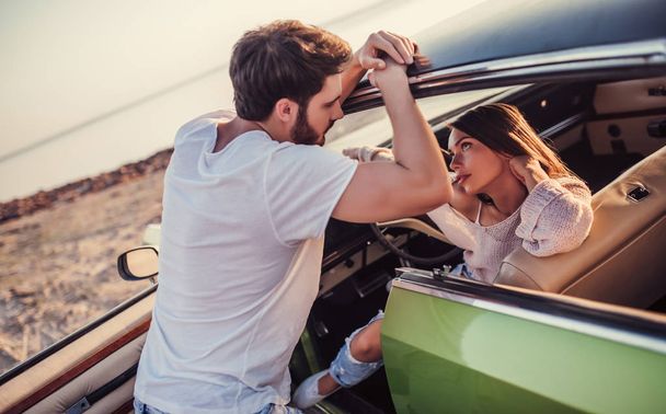 Romantic couple with green retro car on the beach. Handsome bearded man and attractive young woman with vintage classic car. Stylish girl is sitting in car while her man is standing near and is gonna kiss her. Love story. - Foto, Bild