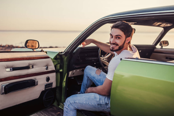 Handsome bearded man is sitting in his green retro car standing on the beach. Vintage classic car. - Foto, Bild