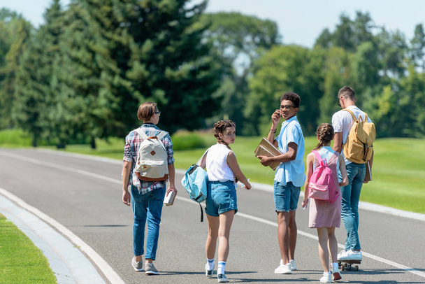 back view of multiethnic teenage friends with books and backpacks walking together in park - Foto, afbeelding