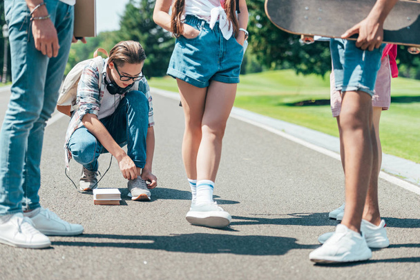 cropped shot of teenagers with books and skateboard waiting for friend tying shoelaces in park - 写真・画像