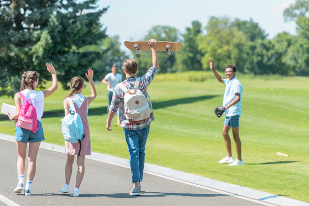 teenagers walking with books and skateboard in park and greeting classmates playing baseball - Fotoğraf, Görsel