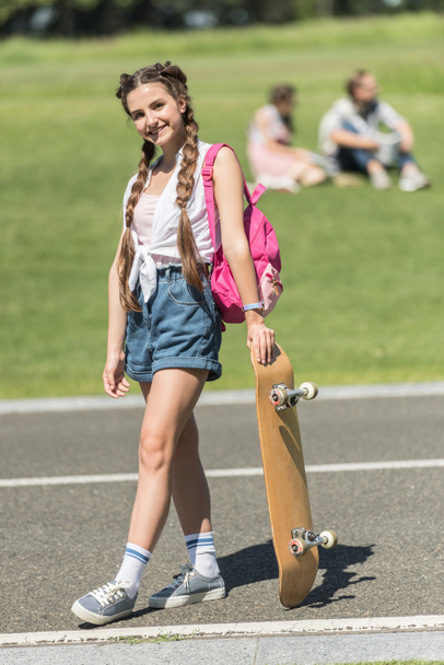 beautiful teenage girl with backpack standing with skateboard and smiling at camera in park - Fotó, kép