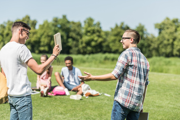 happy teenage boys smiling each other and holding book while friends studying behind in park - Fotoğraf, Görsel