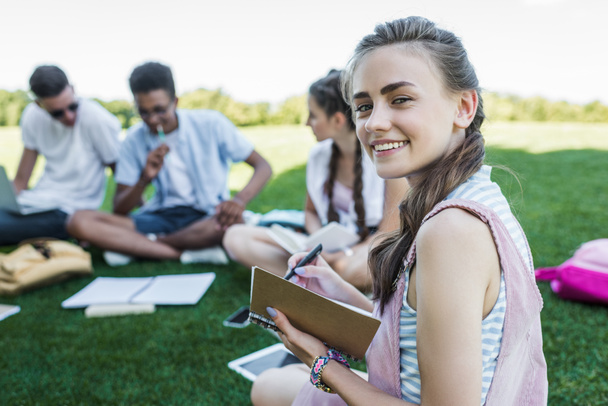 feliz adolescente tomando notas y sonriendo a la cámara mientras estudia con amigos en el parque
 - Foto, Imagen