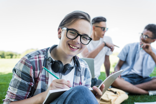 happy teenage boy taking notes and smiling at camera while studying with friends in park - Photo, image