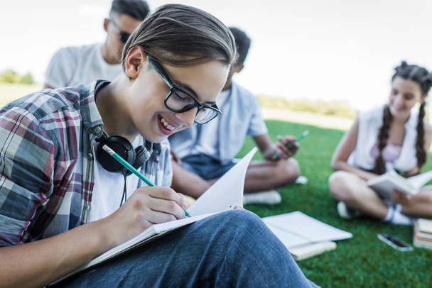 smiling teenage boy writing in notebook while studying with friends in park - Foto, Imagen