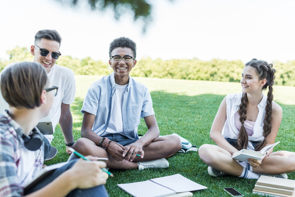 estudiantes adolescentes multiétnicos felices sentados en la hierba y estudiando juntos en el parque
 - Foto, Imagen