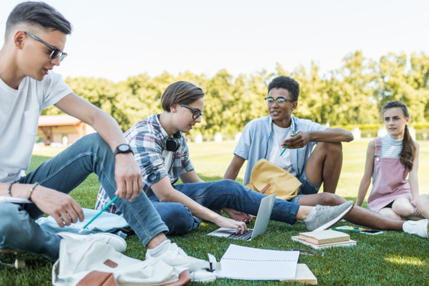 happy multiethnic teenagers sitting and talking while studying together in park  - Foto, afbeelding