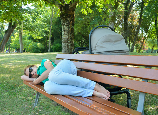 Unavená matka spí na lavičce v parku s dítětem v kočárku - Fotografie, Obrázek