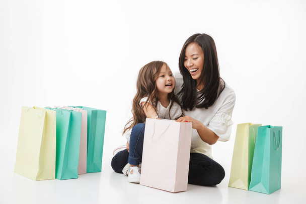 Image of happy young asian woman mother with her little girl child daughter isolated over white wall background with shopping bags. - Foto, afbeelding