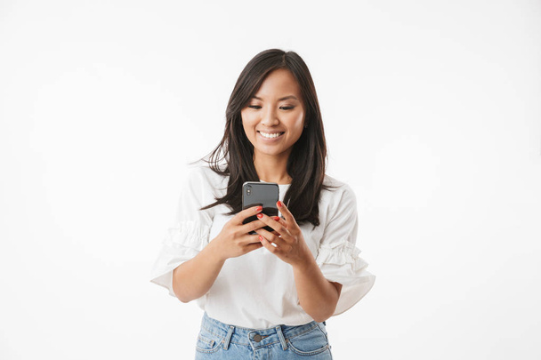Image of satisfied asian woman with long dark hair smiling and looking at cell phone holding in hand isolated over white background - Photo, Image