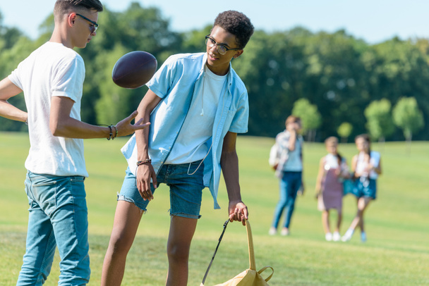 adolescentes amigos multiétnicos jugando con pelota de rugby mientras sus compañeros de clase caminan detrás en el parque
  - Foto, Imagen