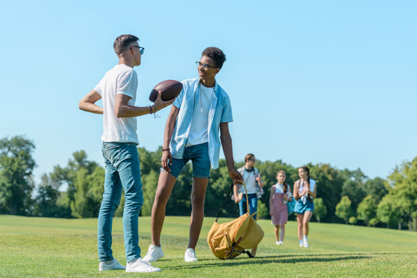 chicos multiétnicos jugando con pelota de rugby mientras sus compañeros de clase caminan detrás en el parque
  - Foto, imagen
