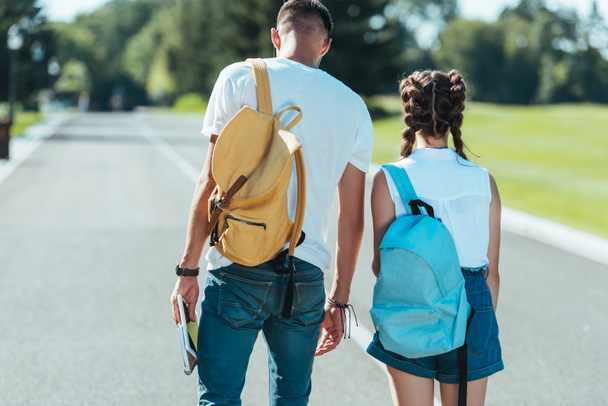 Rückansicht eines Jungen und eines Mädchens mit Rucksäcken, die gemeinsam im Park spazieren - Foto, Bild