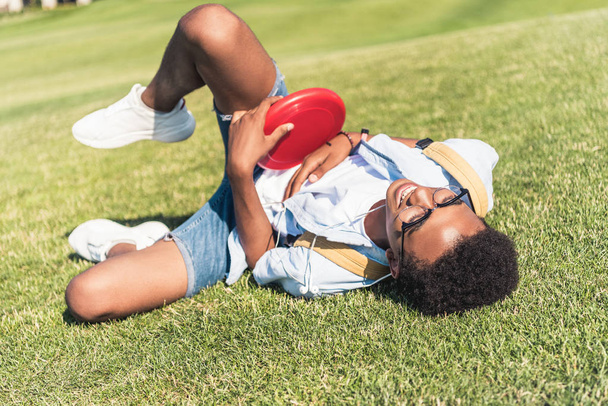 happy african american teenager holding lying disk and lying on grass in park - 写真・画像