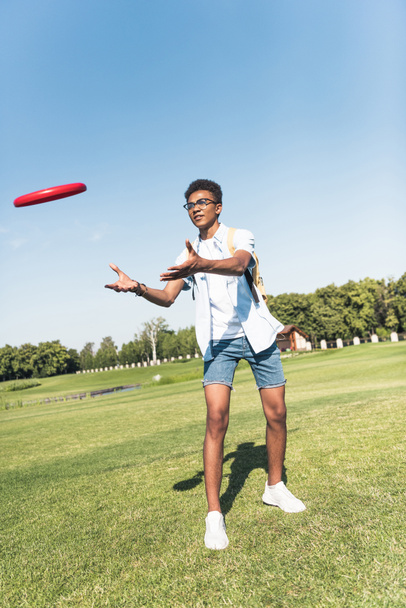 full length view of african american teenager with backpack playing with flying disc in park - Fotografie, Obrázek