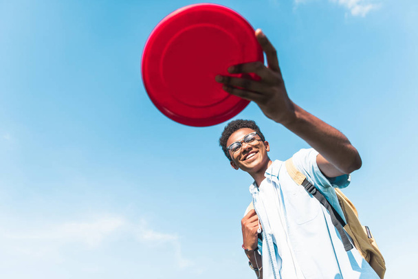 low angle view of happy african american teenager holding flying disc against blue sky - Zdjęcie, obraz