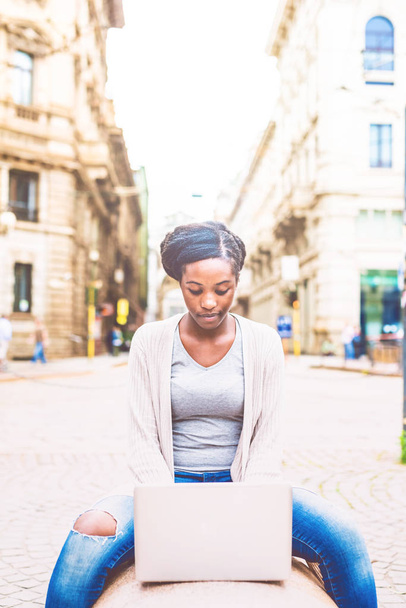 Giovane donna africana che utilizza il computer all'aperto in città seduta su una panchina toccando la tastiera
 - Foto, immagini