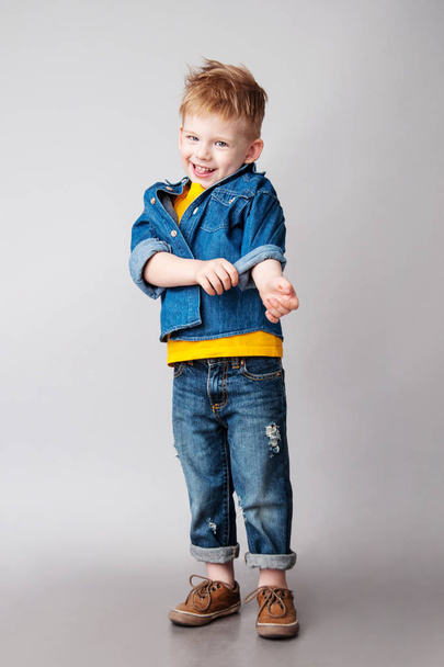 Porträt eines modernen modischen Jungen, der vor grauem Hintergrund posiert. Bekleidet ist er mit einem gelben T-Shirt, Jeansjacke, blauen Jeans und braunen Turnschuhen. Studioaufnahme  - Foto, Bild