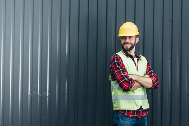 male worker in safety vest and helmet standing with crossed arms near wall - Фото, изображение