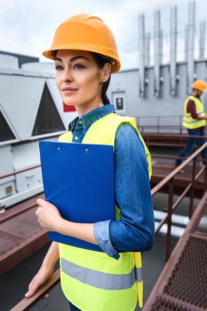 female architect in helmet holding clipboard on roof, male colleague behind - Photo, Image