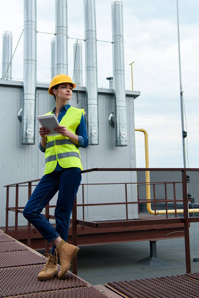 female architect in safety vest and hardhat using digital tablet on industrial construction - Fotó, kép