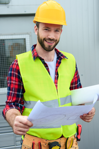 smiling engineer in safety vest and helmet working with blueprints - Φωτογραφία, εικόνα