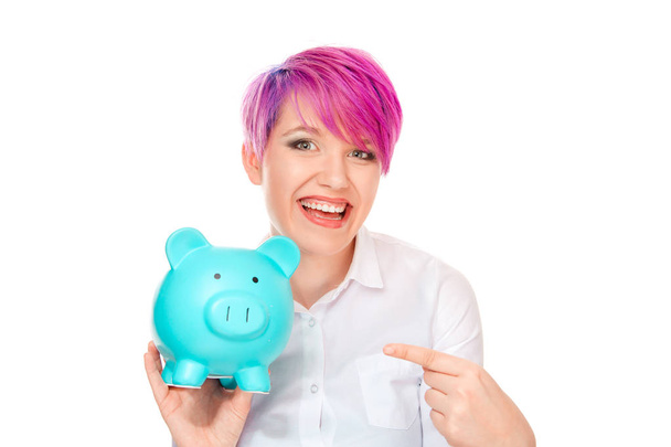 Confident stylish woman with pink hair holding blue piggy bank and pointing at it with happy smile looking at camera isolated on white background - Foto, Imagen