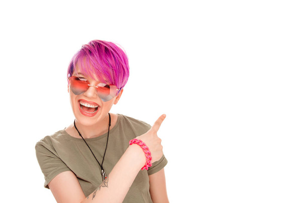 Cheerful vivid woman with pink hair wearing sunglasses and pointing behind back while smiling at camera on white background - Photo, image
