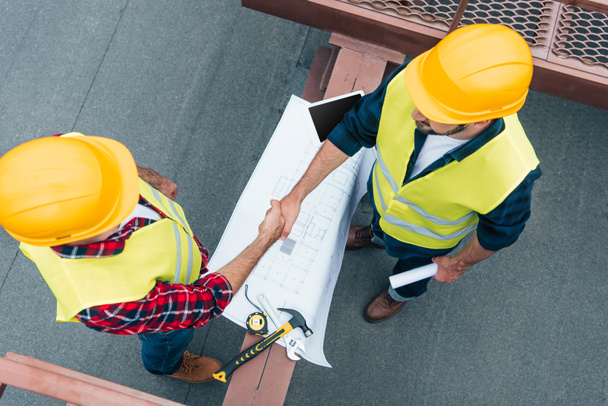 overhead view of engineers in helmets with blueprints shaking hands on roof - Photo, Image