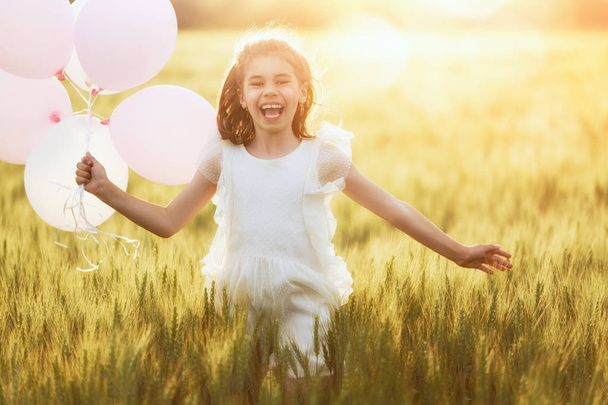 Happy kid is having fun on nature in the summer. Child is laughing and playing on meadow at sunset background. Girl with air balloons is running on cereal field. - Foto, Bild