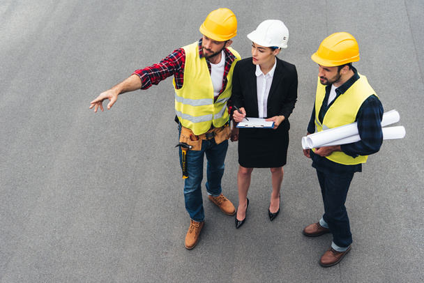 Vista aérea de la ingeniera y los trabajadores masculinos con portapapeles y planos apuntando a la construcción
 - Foto, Imagen