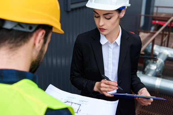 selective focus of female architect and male worker in helmets with blueprint and clipboard on construction - Photo, Image