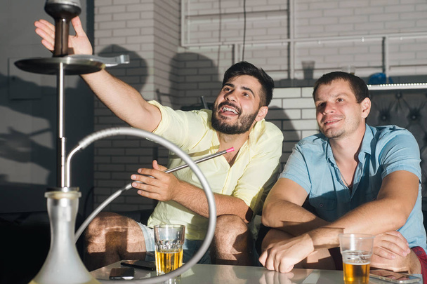Friends men drink beer, smoke a hookah and have fun sitting at the table - Fotoğraf, Görsel