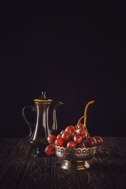 close-up shot of red grapes in vintage metal bowl and teapot on wooden table on black - Fotoğraf, Görsel