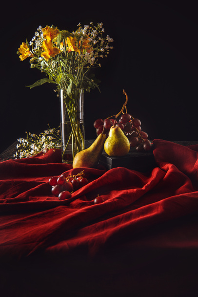 still life with ripe fruits and flowers in vase on red drapery on black - Foto, Bild