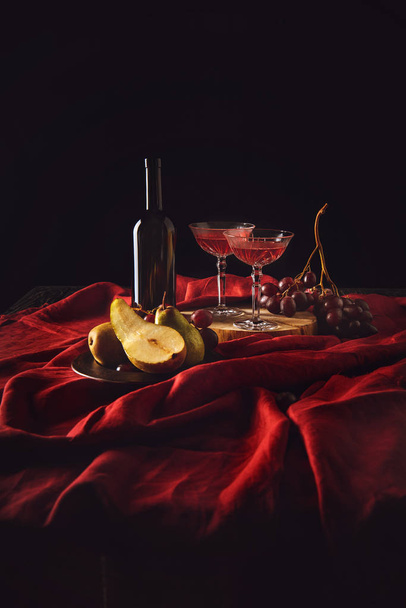 still life with pears, grapes and wine in bottle and crystal glasses on red drapery on black - Photo, Image