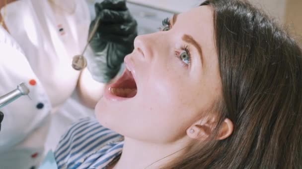 close-up of a cropped frame of a young beautiful girls face at the dentist. The patient lies in the dental chair with an open mouth, the doctor in gloves drills the tooth with a drill and looks at - Кадры, видео