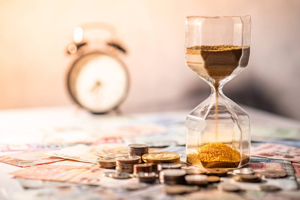 Sand running through the shape of hourglass on table with banknotes and coins of international currency. Time investment and retirement saving. Urgency countdown timer for business deadline concept - Foto, Bild