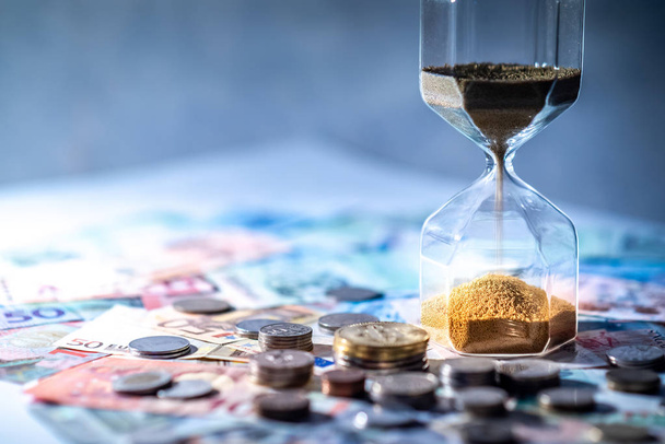 Sand running through the shape of hourglass on table with banknotes and coins of international currency. Time investment and retirement saving. Urgency countdown timer for business deadline concept - Foto, Imagen