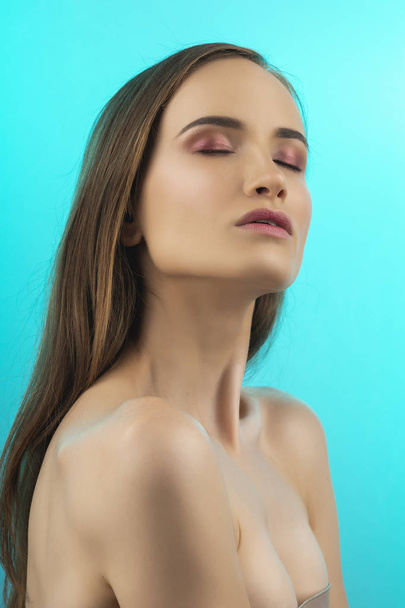 A beautiful girl with naked shoulders and nude makeup closed her eyes with pleasure. Close up. Isolated on blue. Copy space. Advertising and conceptual design. Healthy skin. - Fotografie, Obrázek