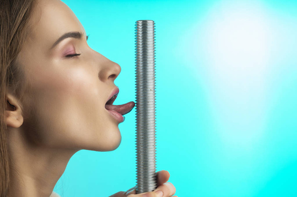 A beautiful girl with naked shoulders and closed eyes touches the thread of big steel bolt with her tongue. Close up. Isolated on blue. Copy space. Advertising and conceptual design. Healthy skin. - Foto, Imagen