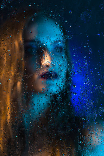 Art style portrait in blue tones of a lonely beautiful sad model girl behind a window glass, over which rain drops down. Artistic blur on model face. Conceptual, commercial and fashionable. Copy space - Valokuva, kuva