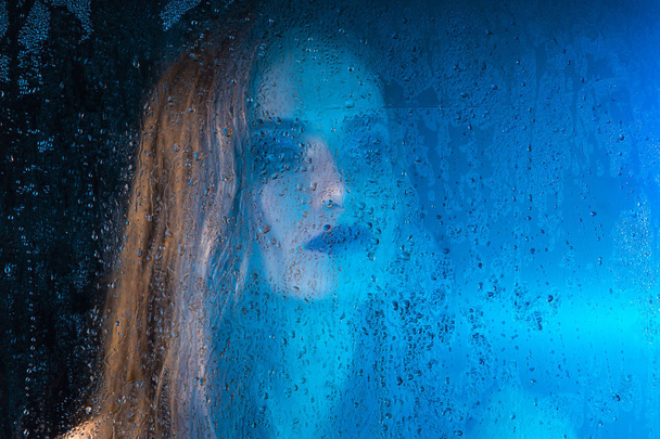 Art style portrait in blue tones of a lonely beautiful sad model girl behind a window glass, over which rain drops down. Artistic blur on model face. Conceptual, commercial and fashionable. Copy space - Фото, зображення