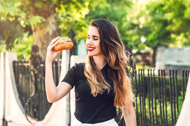 девушка смотрит на бургер. Мясо посередине с соусом. празднование. длинные волосы. улыбка на лице - Фото, изображение
