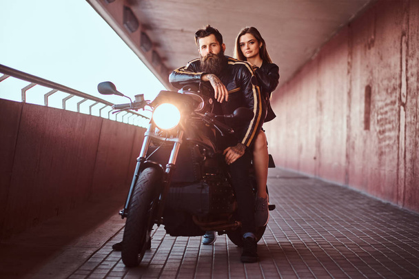 Brutal bebaarde biker in zwart lederen jasje en sensuele brunette meisje zitten samen op een op maat gemaakte retro motorfiets op een wandelpad onder een brug. - Foto, afbeelding