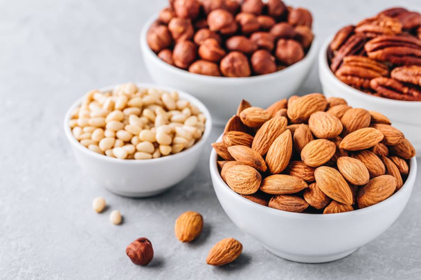 Almonds, pecans, pine nuts and hazelnuts in white bowls on grey background. Nuts mix. Healthy food and snack. - Photo, Image