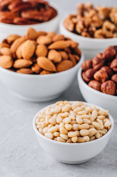 Pine nuts, almonds, pecans, walnuts and hazelnuts in white bowls on grey background. Mixed nuts. Healthy food and snack. - Photo, Image